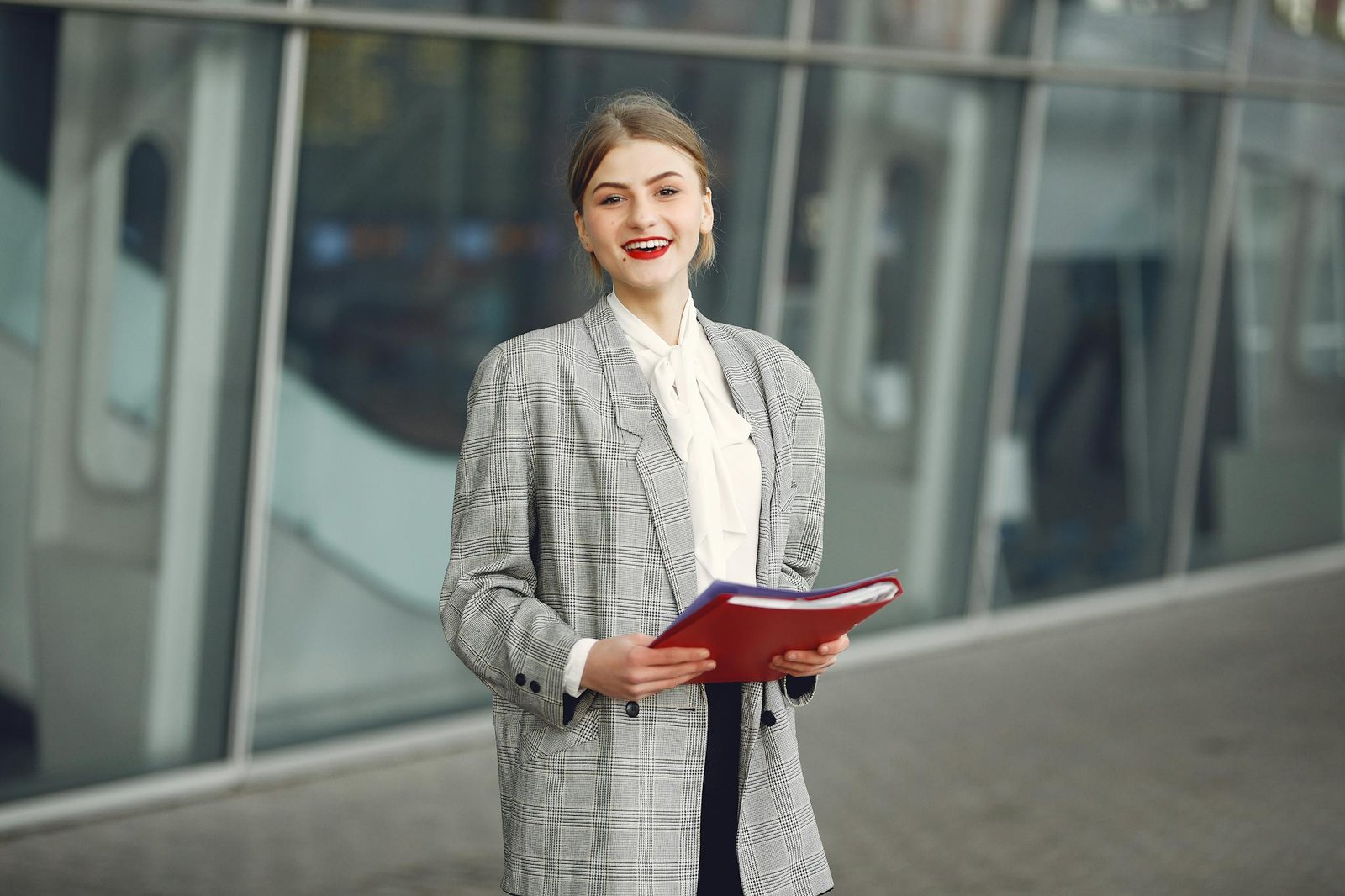 Positive young woman in trendy wear smiling at camera while holding documents standing against glass building in downtown